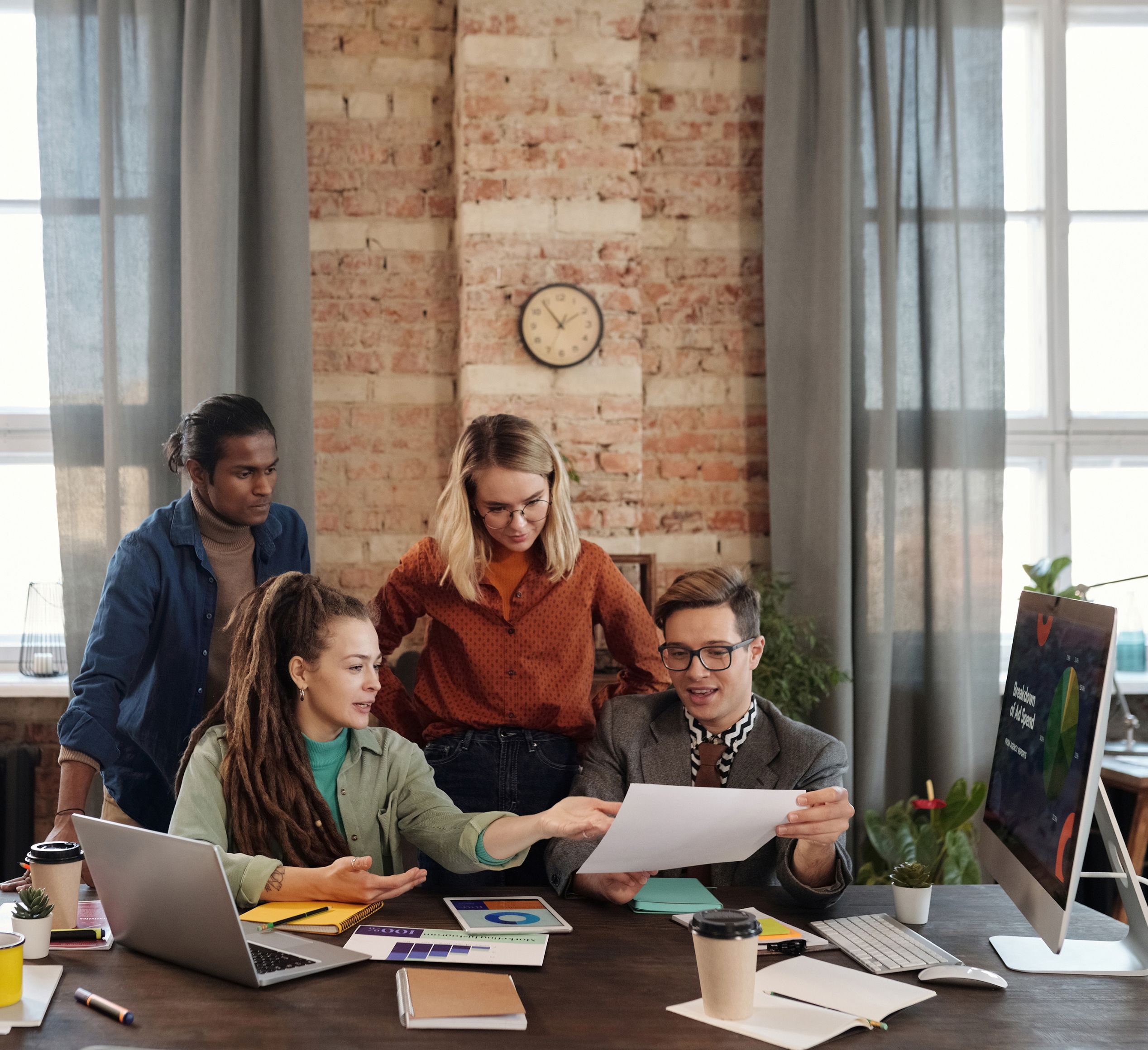 A lifestyle photo showing a small marketing team of four people at a desk, looking at screens and papers. On the left is a woman with dreadlocks and a green shirt sitting at the desk, and there is a man with a ponytail and blue shirt standing behind her. On the right there is a man with short brown hair and glasses holding up a piece of paper, and standing behind him is a woman with blonde hair and glasses in an orange shirt. All look happy, interested and curious.
