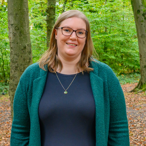 Website profile pic A photo of Becki Wood, Founder of Pyjama Shark Marketing. Becki has dark blonde shoulder length hair, blue eyes and wears glasses. She is standing in a wooded area with trees around her, and she is wearing a dark top with teal green blazer style coat.